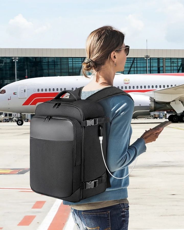 Sac à Dos Cabine Avion 40x20x25, porté sur le dos d'une femme avec son téléphone en main, en train de charger grâce à la prise USB du sac, dans un aéroport