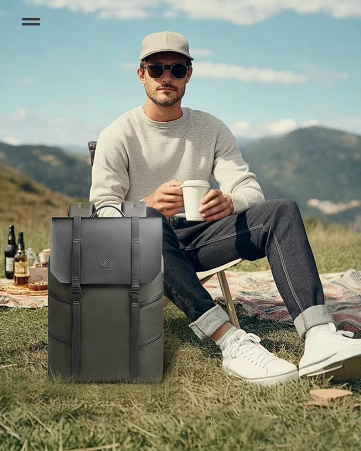 Homme assis en plein air prenant un café avec un sac à dos ordinateur design kaki posé sur l’herbe.